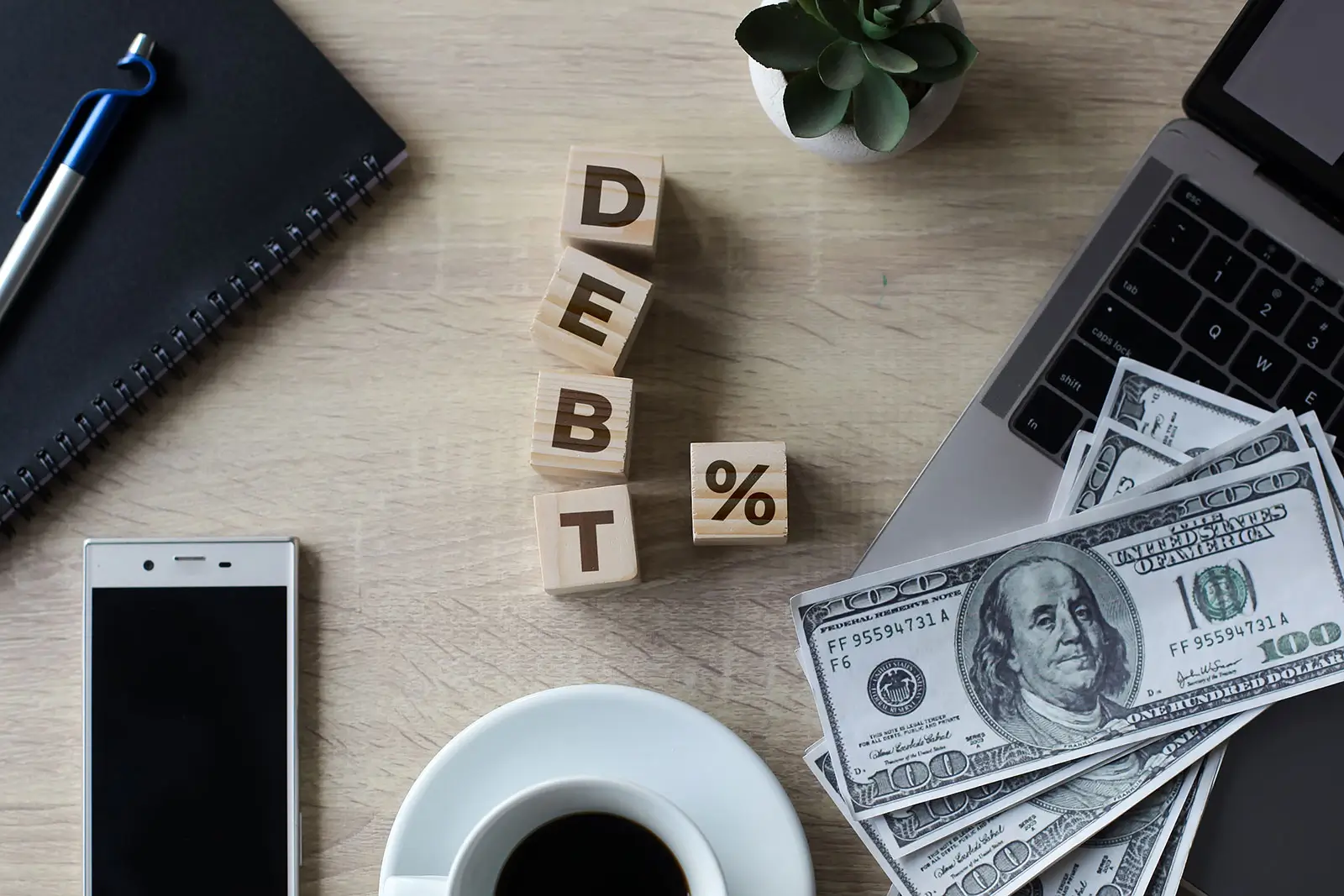 An overhead shot of a desk with wooden blocks spelling "DEBT %" arranged diagonally. Surrounding the blocks are various financial and office items: a black notebook with a blue pen, a white smartphone, a white cup of black coffee, a laptop, a small potted plant, and a stack of US hundred-dollar bills. The image visually represents debt, interest rates, and financial management or challenges.